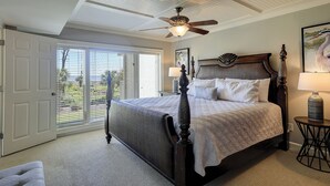 Een slaapkamer met een groot bed, een plafondventilator, een raam met jaloezieën en uitzicht op groen.