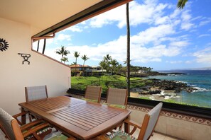 Una mesa y sillas de madera en una habitación con vistas a un paisaje costero, que incluye palmeras y una playa.