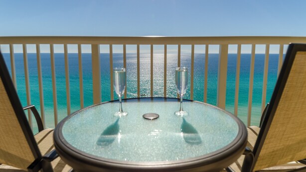 A round glass table with two champagne flutes on a balcony overlooking a turquoise sea. - Paradise Harbor Grand Panama PCB-Beachfront 2Bd+Bunks-2Ba-Pet Friendly (Panama City Beach)