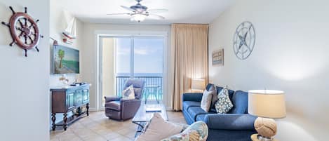 Una sala de estar con un sofá, un sillón y una mesita. Se observa un ventilador de techo, un reloj y una ventana con cortinas.