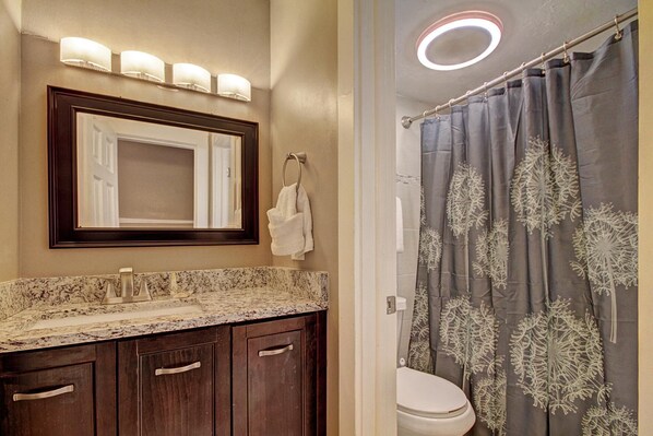 Un baño con lavamanos de mármol, armarios de madera oscura, un espejo con iluminación y una cortina de ducha con motivos de hojas.