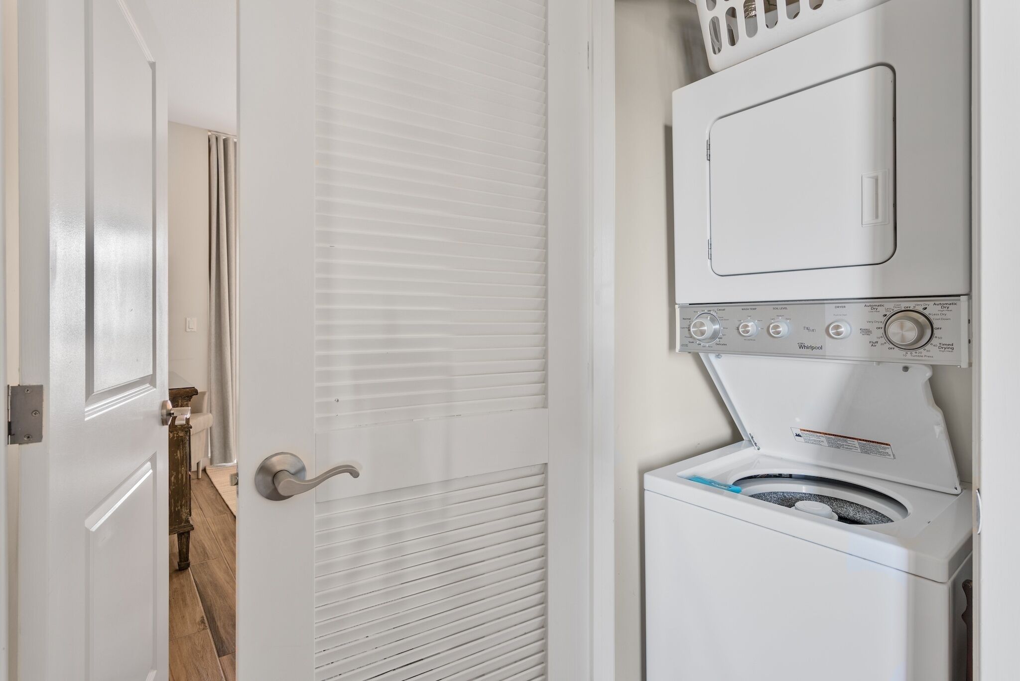 A laundry room with a stacked washer and dryer, a door with a silver handle, and a wooden floor.