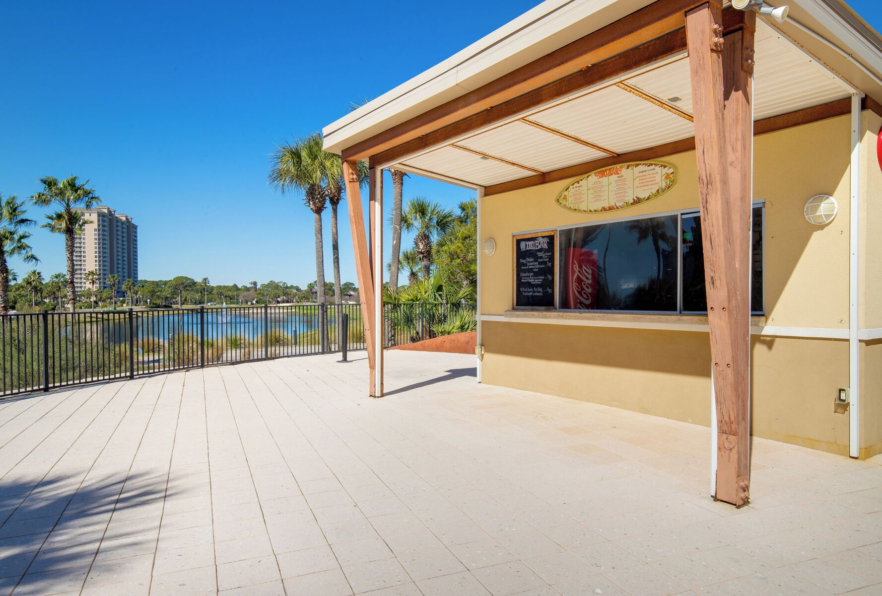 A covered outdoor seating area with a menu board, a view of a body of water, and palm trees.