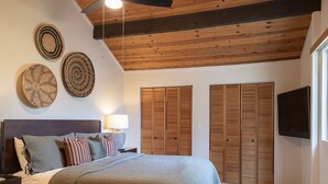 Un dormitorio con cama, mesitas de noche, ventilador de techo, cabecera de madera y televisor montado en la pared.