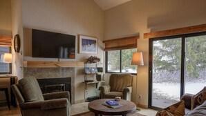 Una sala de estar con chimenea, asientos cómodos, un televisor de pantalla plana y ventanas amplias con vistas al exterior.