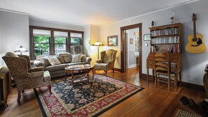 A living room with a sofa, armchairs, a coffee table, a rug, a bookshelf, a guitar, and a window with a view of greenery. - Beautiful Cheyenne Cañon Retreat with Hot Tub, Walk To Broadmoor Hotel & Trails. (Colorado Springs)