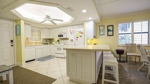 Eine Küche mit weißen Schränken, einer Mikrowelle, einem Kühlschrank und einem Deckenventilator. Es gibt eine Essecke mit Tisch und Stühlen.