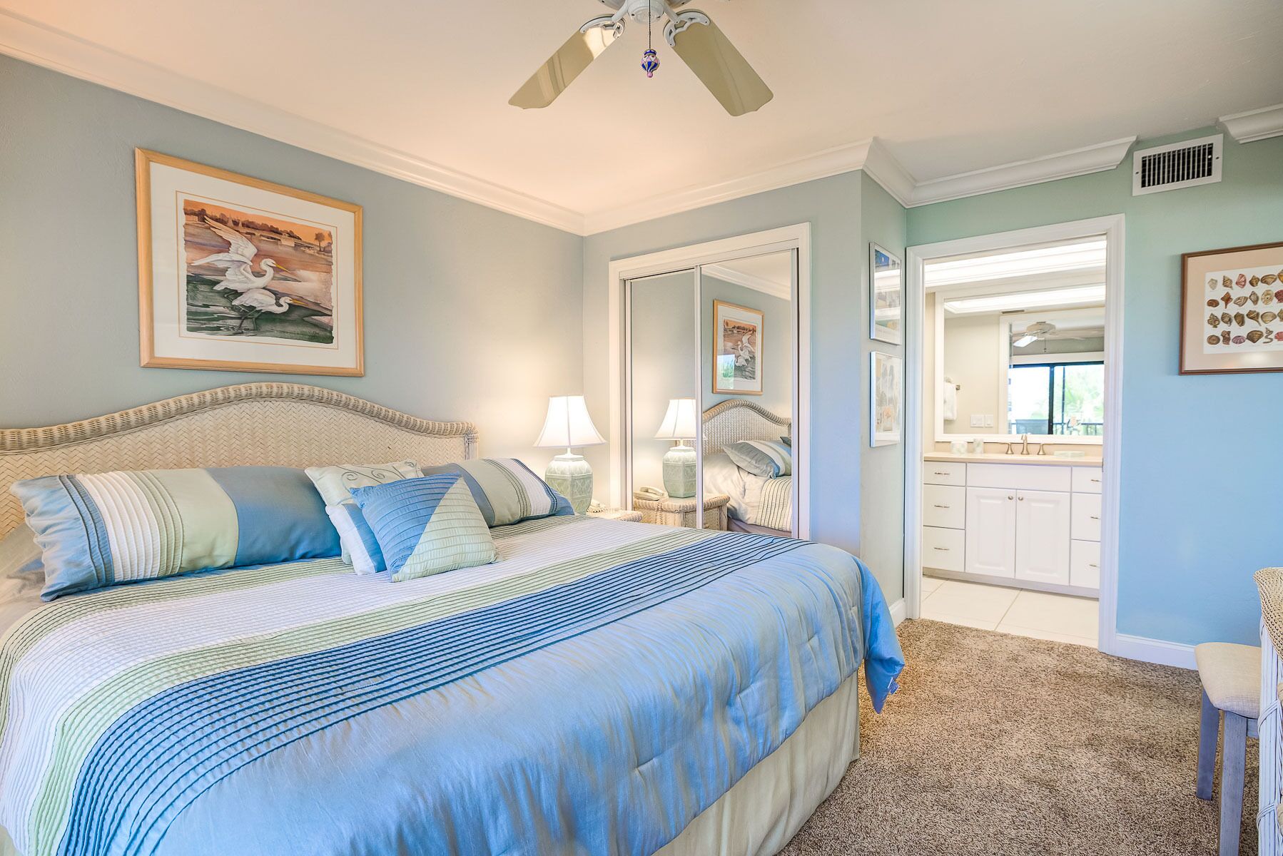 A bedroom with a large bed, a ceiling fan, a bathroom with a vanity, and a door leading to another room.