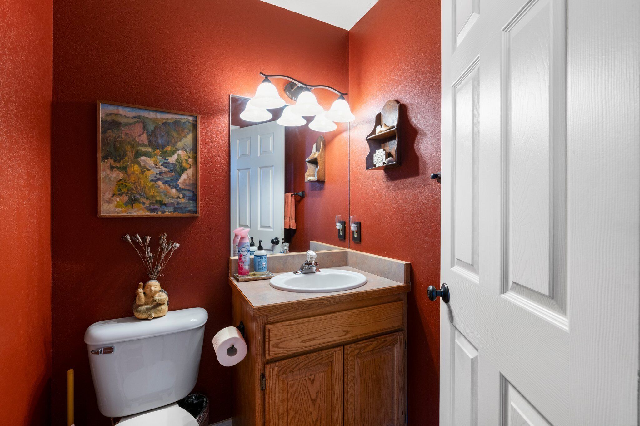 A bathroom with a wooden vanity, a white toilet, a mirror with lights, a painting on the wall, and a door.