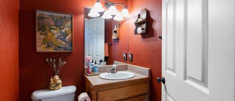 A bathroom with a wooden vanity, a white toilet, a mirror with lights, a painting on the wall, and a door.