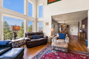 Relax in this stylish living room with a high ceiling and large windows offering a view of the surrounding landscape. The room features a sofa, armchair, and ottoman, along with a coffee table and side table with a lamp.