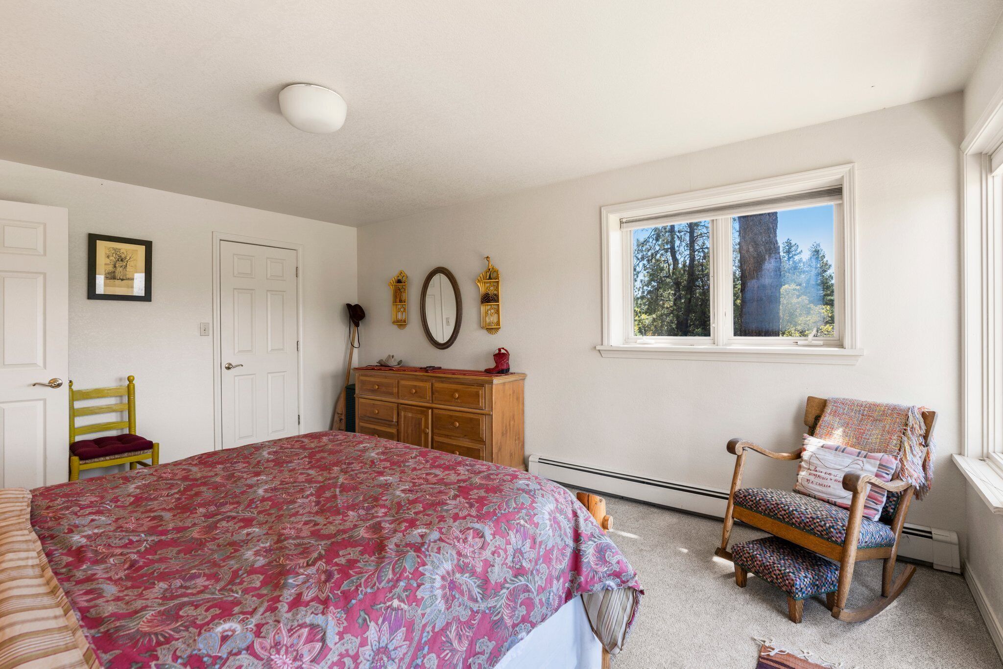A bedroom with a large bed, a wooden dresser, a rocking chair, and a window with a view of trees.