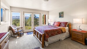 Een slaapkamer met een groot bed, een schommelstoel, een nachtkastje en uitzicht op bergen door de ramen.