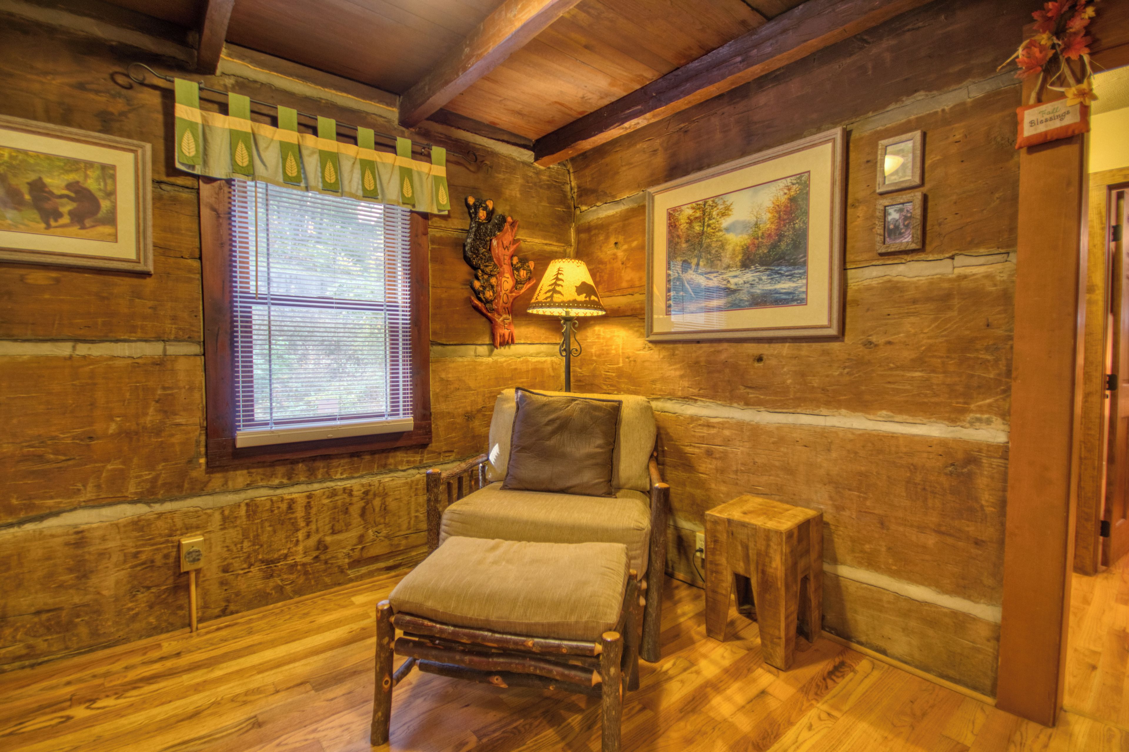 A cozy room with wooden walls, a window with blinds, a chair, a small table, and a lamp.