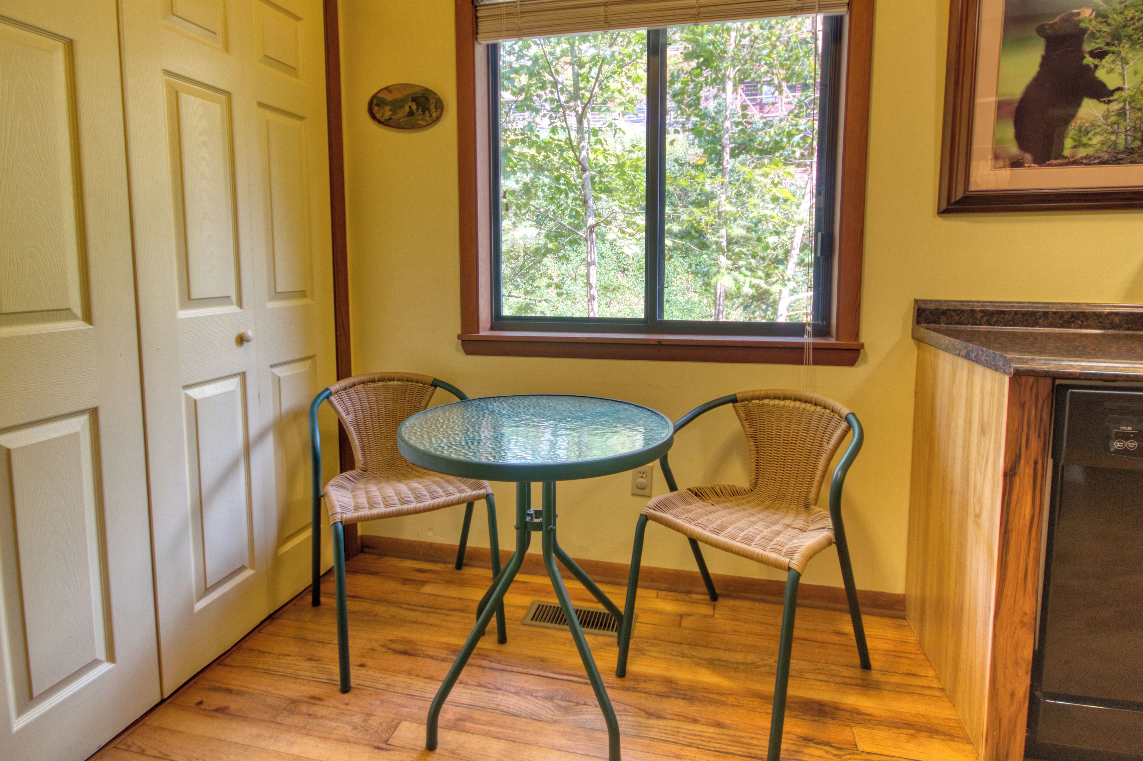 Une pièce avec une table ronde et deux chaises en osier, une fenêtre donnant sur des arbres et une porte avec un interrupteur sur le mur.