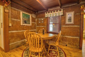 A wooden dining room with a round table and four chairs, framed pictures on the walls, and a window with blinds. - Historic Riverside Cabin w/Hot Tub in Gatinburg (GATLINBURG)