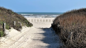 Ein sandiger Strandweg führt zum Meer, gesäumt von Vegetation und Holzbalken.