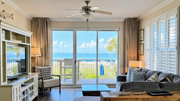 Een woonkamer met een televisie, bank en salontafel, een plafondventilator en een balkon met uitzicht op zee.
