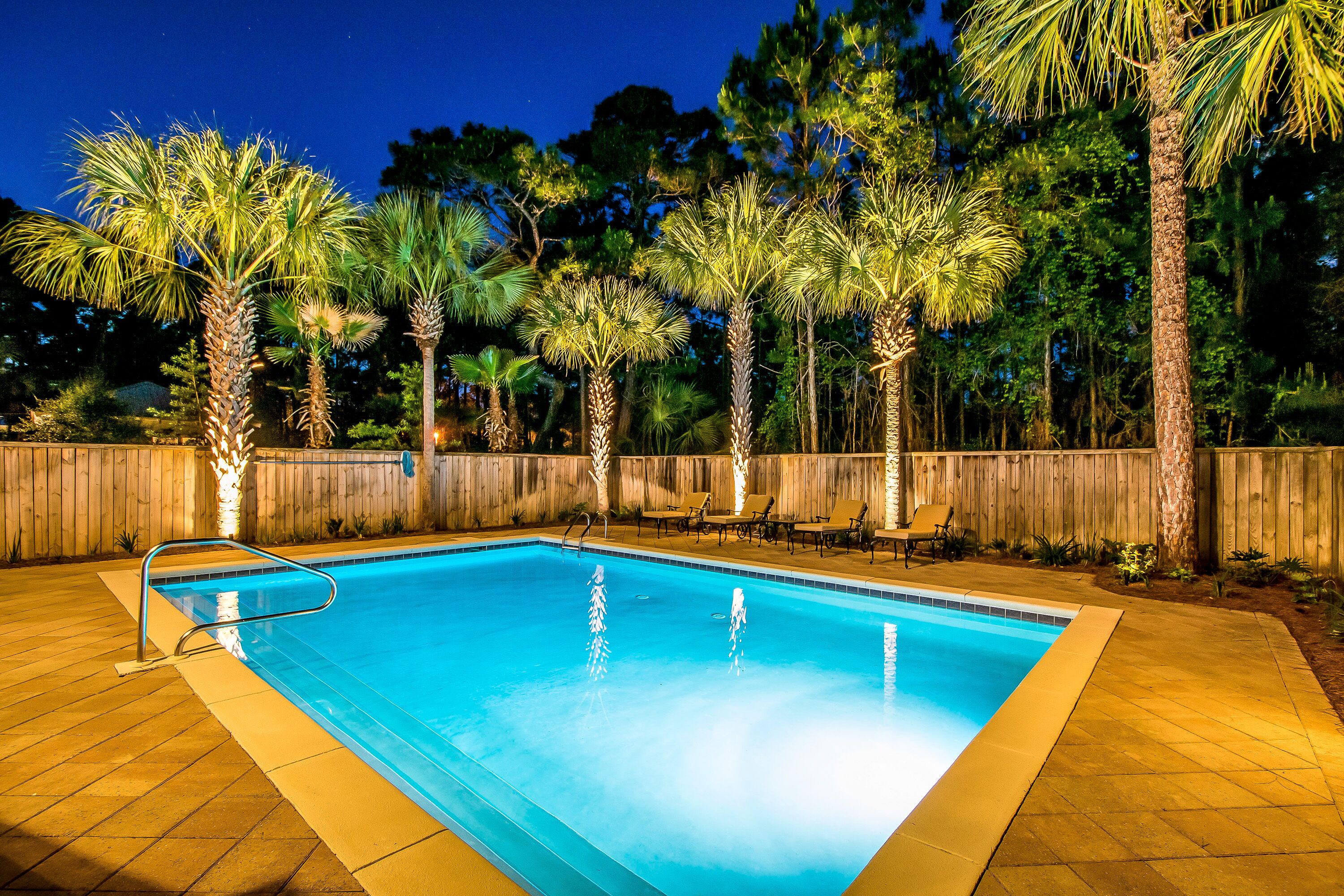 A rectangular swimming pool with a metal ladder on one side, surrounded by palm trees and a wooden fence.