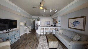 A modern living room with a flat-screen TV, a dining area with bar stools, a kitchen with a bar, and a ceiling fan. - Beautiful Condo! Free round of Golf & Activities Every Day! (Panama City Beach)