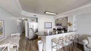 A modern kitchen with a central island, bar stools, and a dining area with a white table and chairs. - Beautiful Condo! Free round of Golf & Activities Every Day! (Panama City Beach)