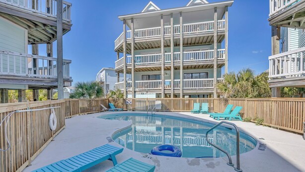 A pool area with blue lounge chairs, a wooden fence, and a multi-story building with balconies.