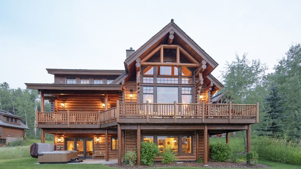 Une grande cabane en rondins dotée d’un balcon, d’un espace extérieur aménagé et couvert, et d’une pelouse bien entretenue.