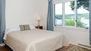 A bedroom with a large bed, a nightstand with a lamp, a window with a view of greenery and water, and a patterned rug. - Fun in the Sun - Walk Into Town on Keuka's East Side (Penn Yan)