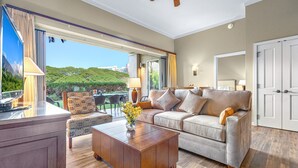 A spacious living room with a large window, a sofa set, a wooden coffee table, and a dining area. - Maui Resort Rentals: Honua Kai Konea 136 – Ground Floor 1BR w/ Lawn Area + Easy Pools/Beach Access (Lahaina)