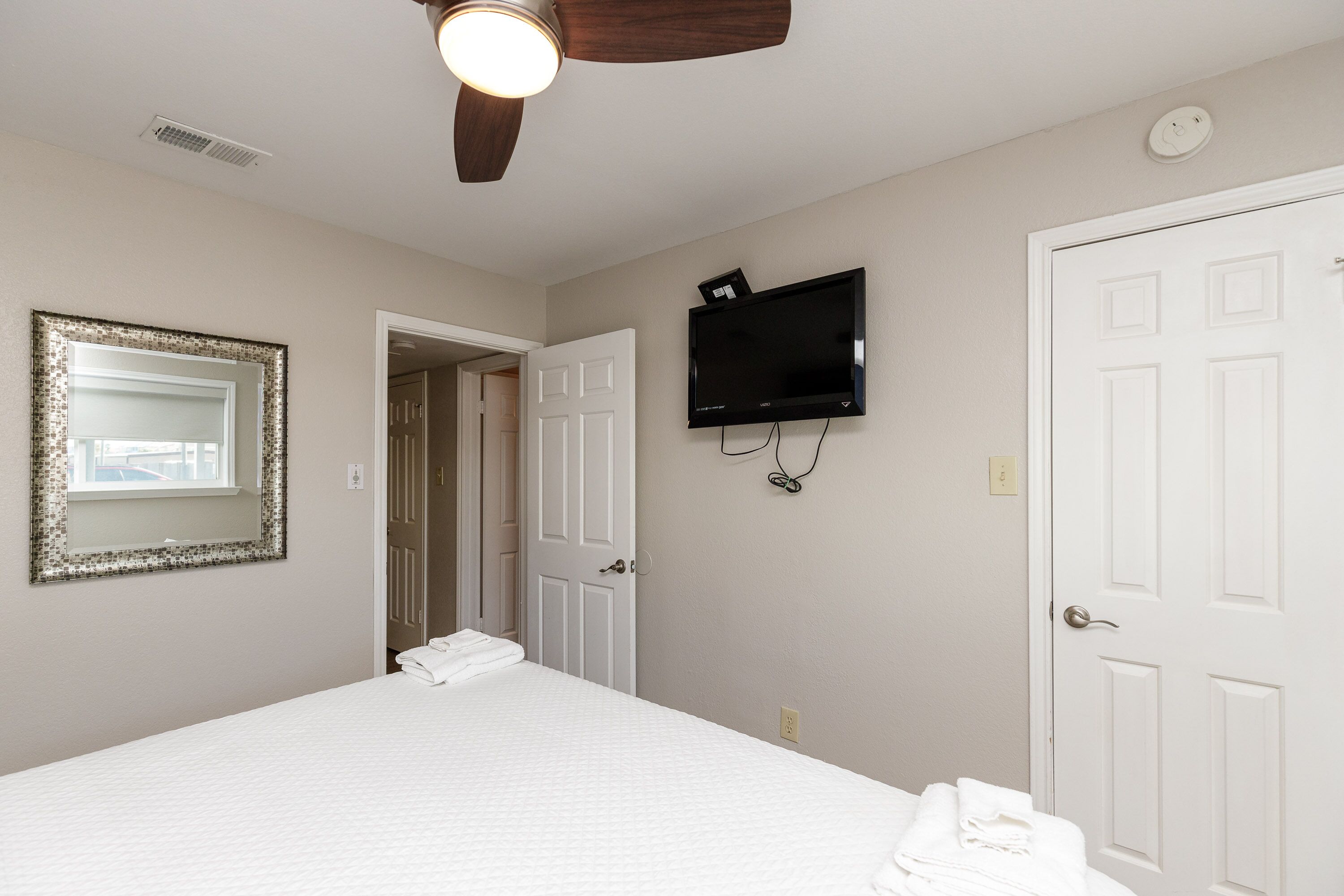 Une chambre à coucher avec un lit, un ventilateur de plafond, un téléviseur fixé au mur et une porte.
