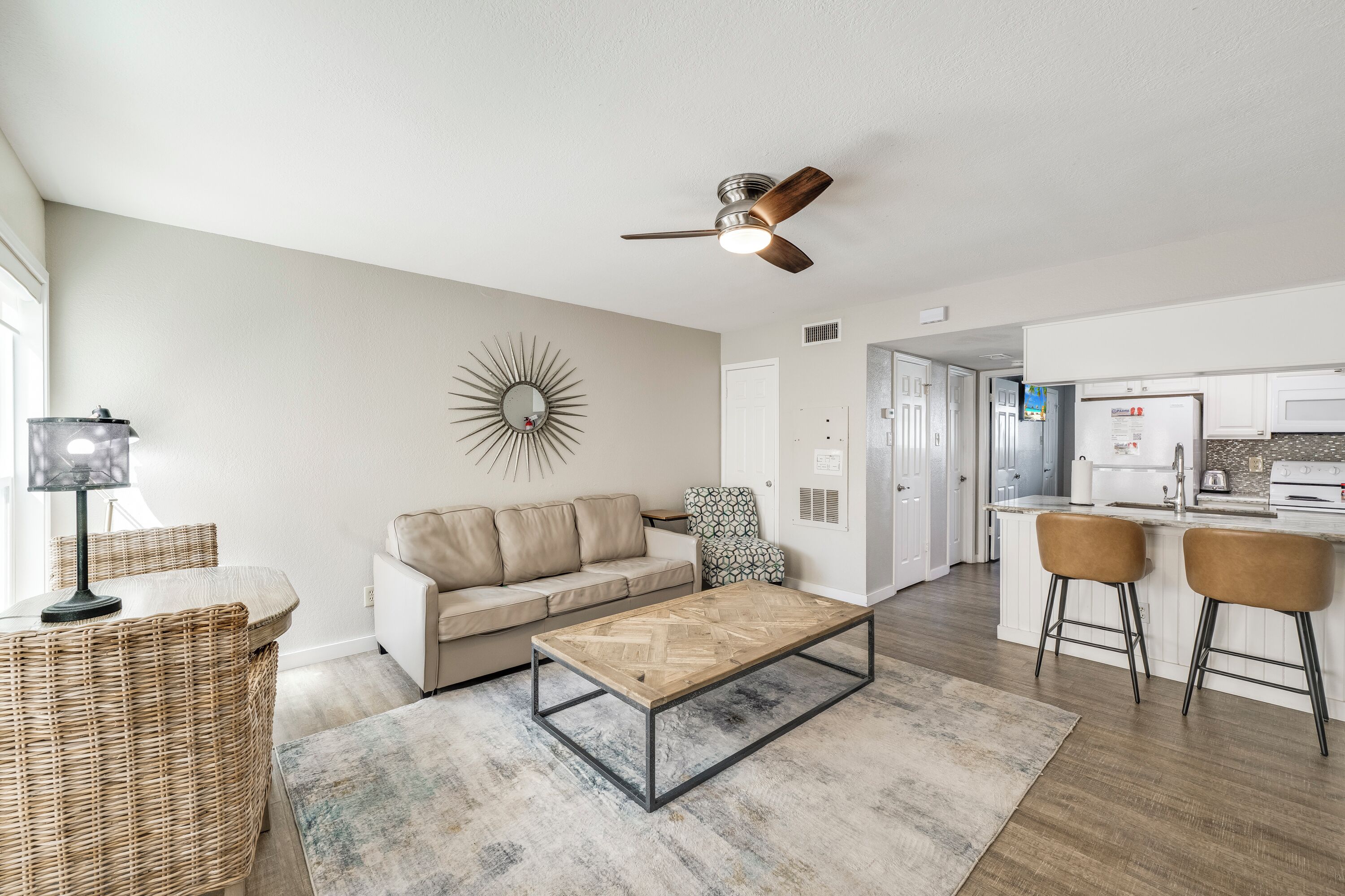 A modern living room with a sofa, coffee table, and chairs, a dining area with bar stools, and a kitchen in the background.