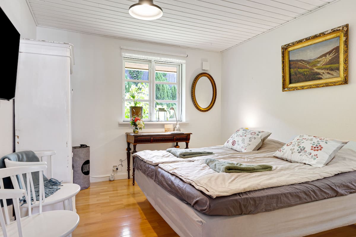 Un dormitorio con cama, una silla, un escritorio, una ventana y un cuadro en la pared.