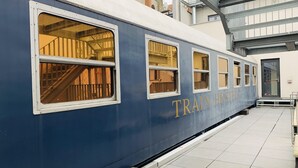 Un vagón de tren azul con la leyenda 'TRAIN HOSTEL', con varias ventanas y una escalera visible a través de una de ellas.