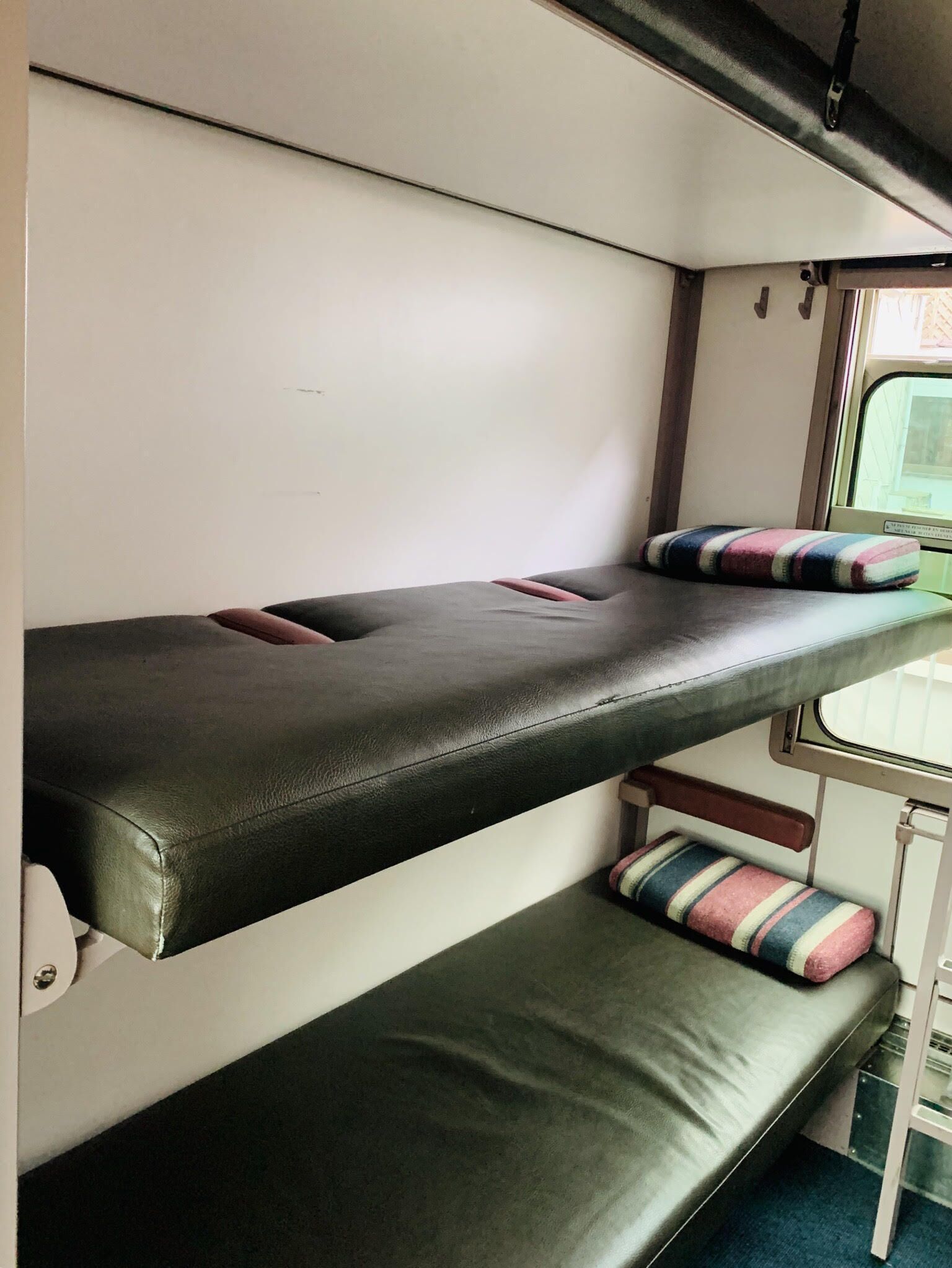 Una cama litera con colchón y almohadas, una ventana con vistas, y una mesita con una silla.
