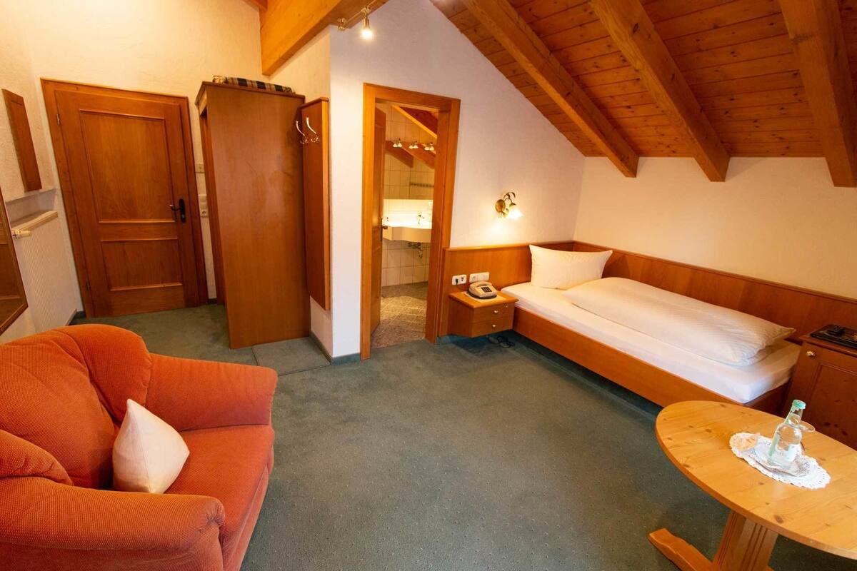 Ein gemütliches Zimmer mit einem Bett, einem Holzschrank, einem kleinen Tisch mit einer Flasche und einer Couch mit Kissen.