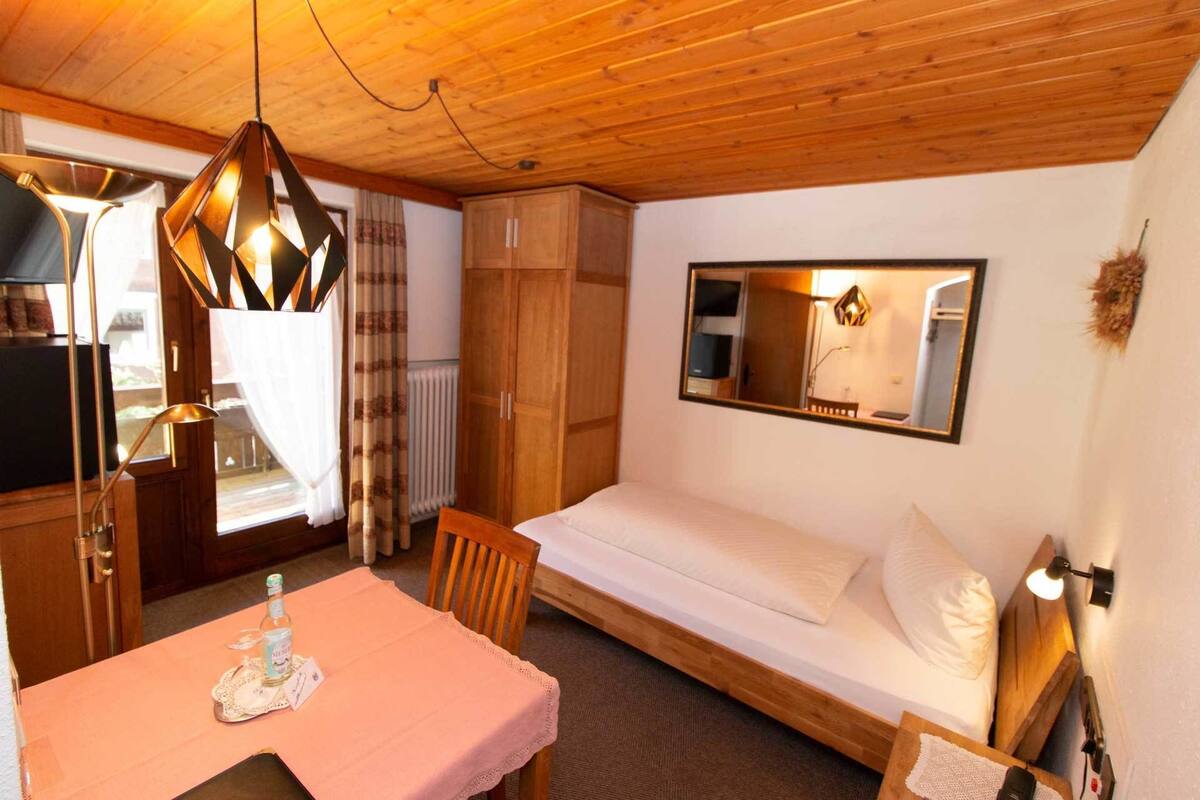 Ein gemütliches Zimmer mit Holzboden, einem Bett mit weißer Bettwäsche, einem kleinen Tisch mit einer rosa Tischdecke und einem Spiegel an der Wand.
