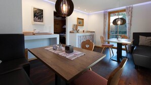 Un coin repas chaleureux avec une table en bois, des chaises et une suspension. On aperçoit une cuisine à l’arrière-plan, avec une fenêtre et des rideaux.