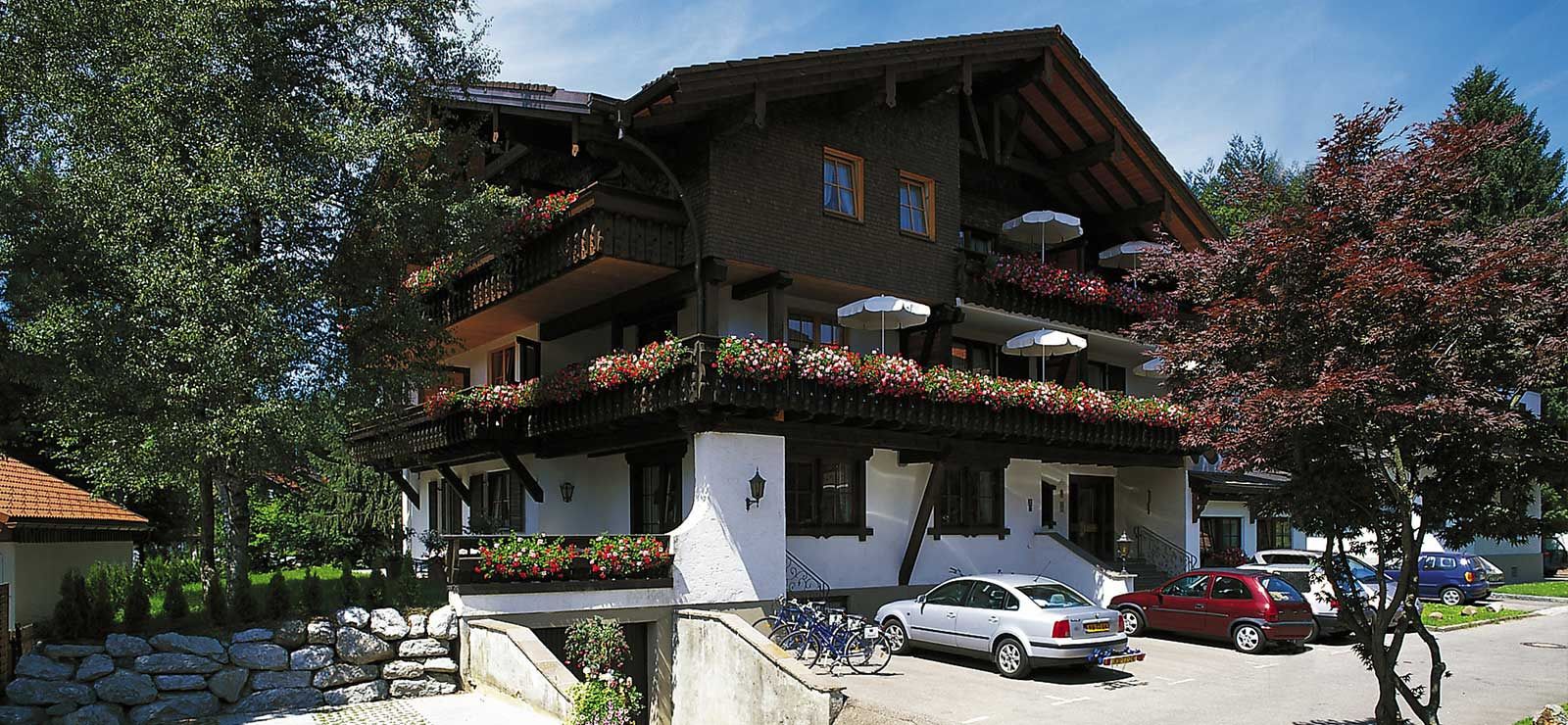 Ein traditionelles Chalet mit Balkonen, die mit Blumen geschmückt sind, einem Kiesweg und geparkten Autos.