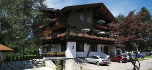 A traditional chalet with balconies adorned with flowers, a stone pathway, and parked cars.