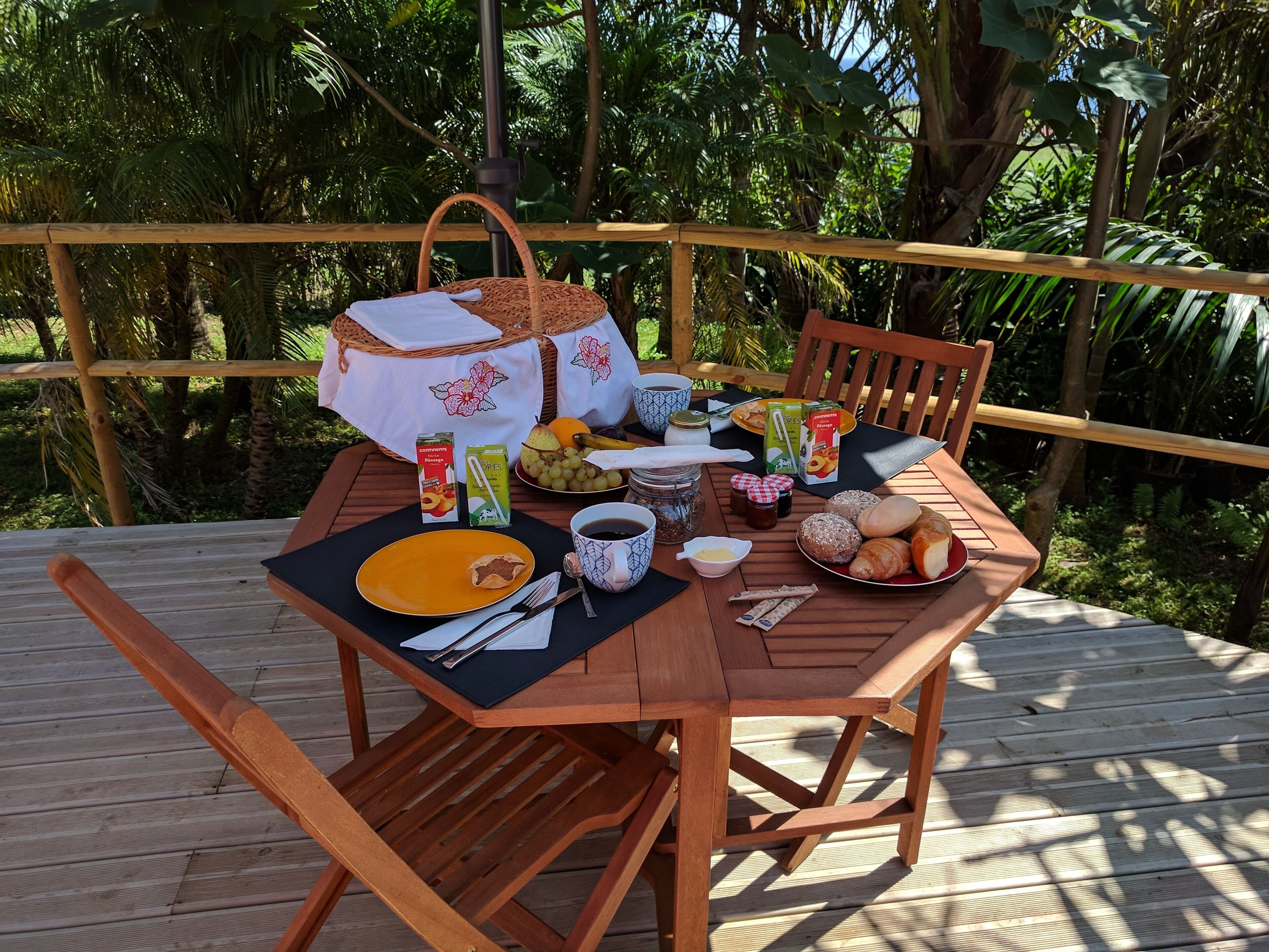 A wooden deck with a table set for breakfast, including bread, fruit, and beverages.