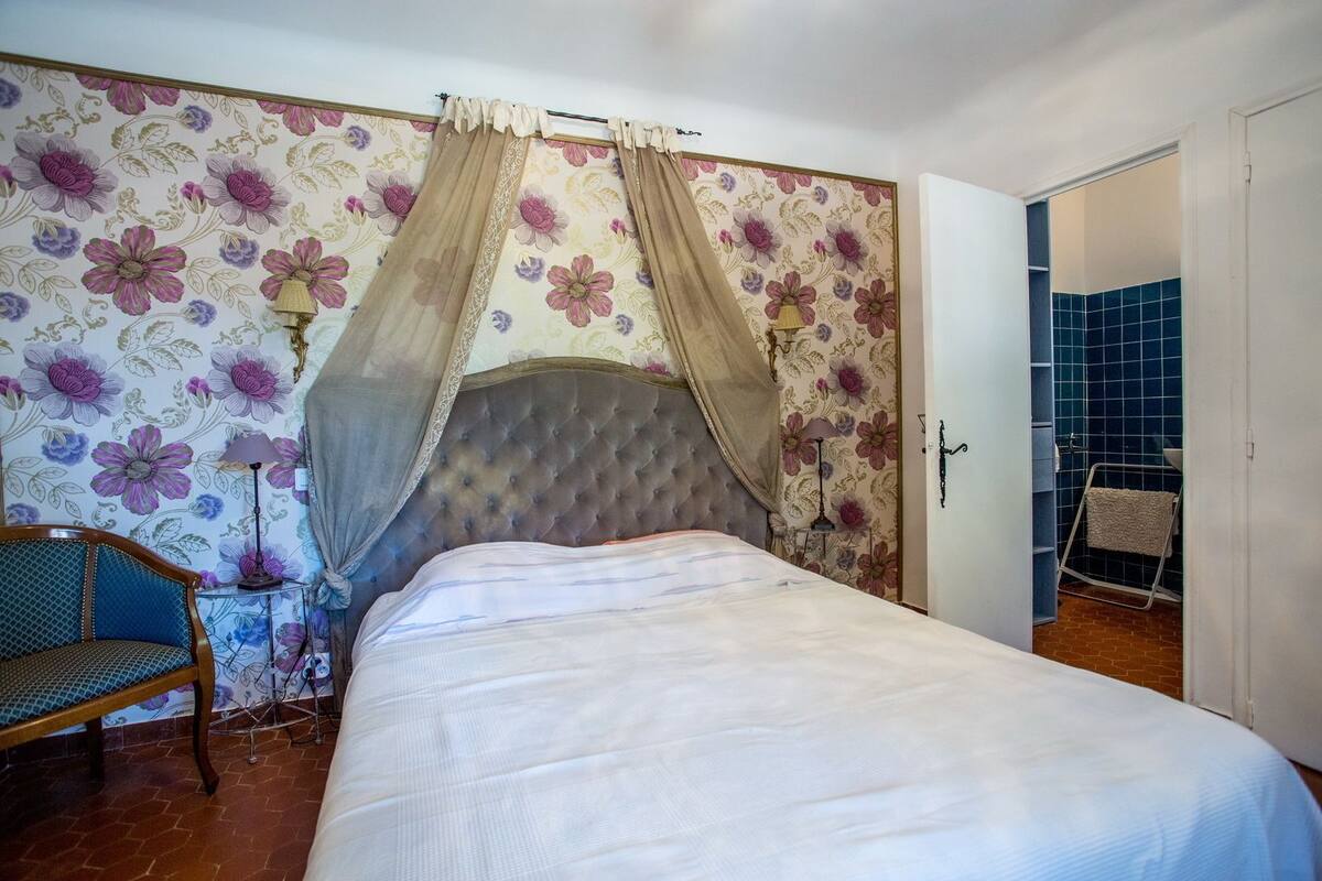 A bedroom with a bed, a chair, a lamp, and a bathroom visible through an open door.