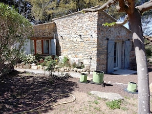 A stone building with a window and a door, surrounded by a garden with plants and a hose. - Domaine l'Helion (Taradeau)