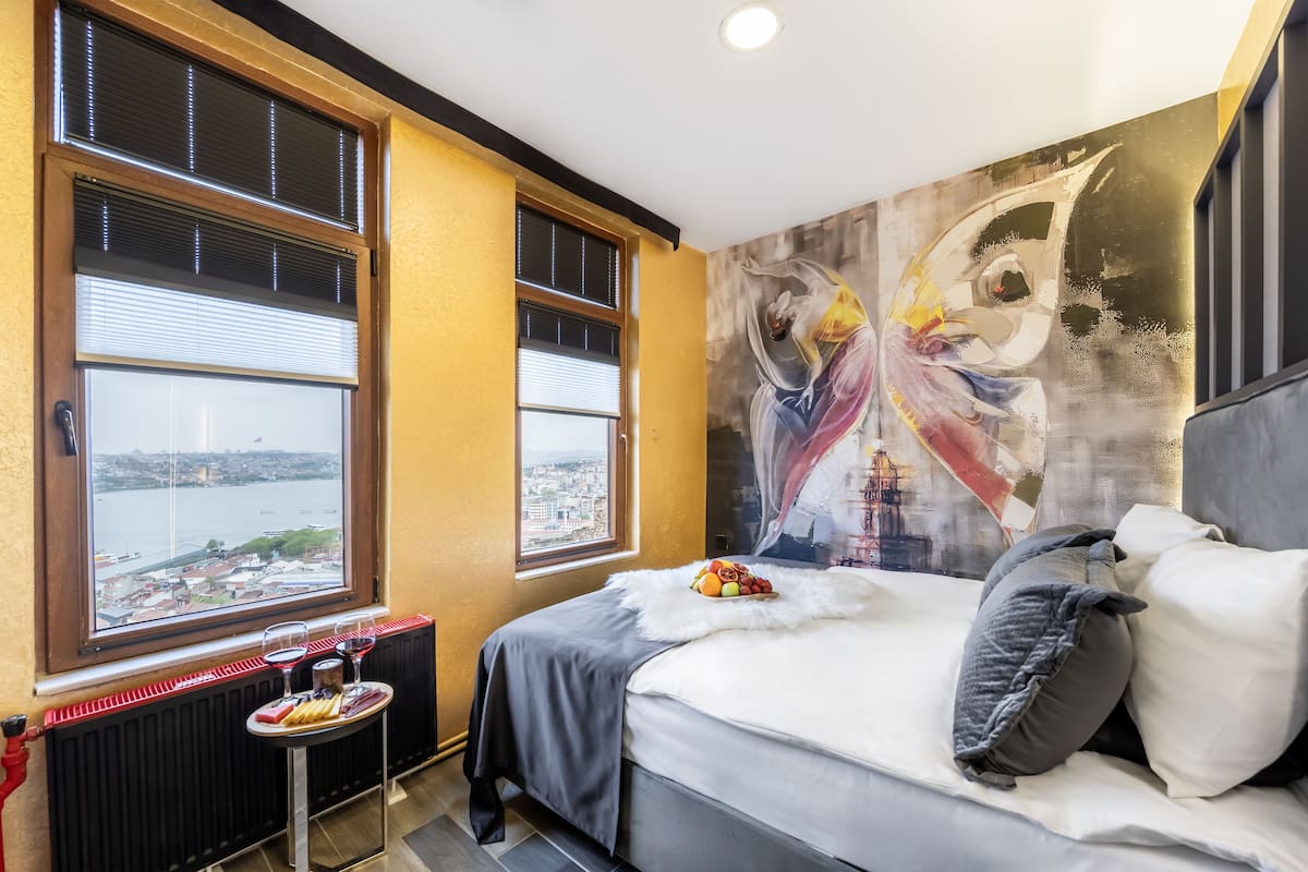 A bedroom with a bed, a window with a city view, and a painting on the wall.
