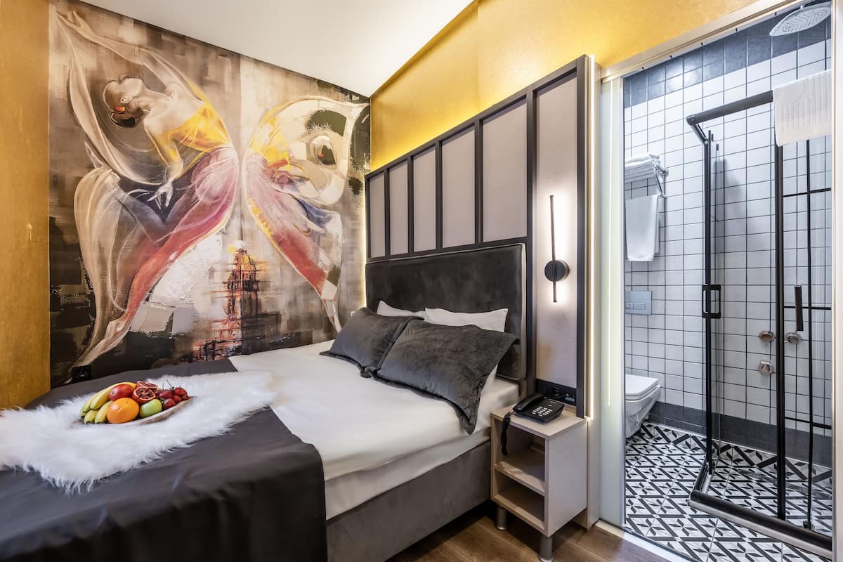 A modern bedroom with a large abstract painting, a neatly made bed with a fruit bowl, and a walk-in shower with patterned tiles.