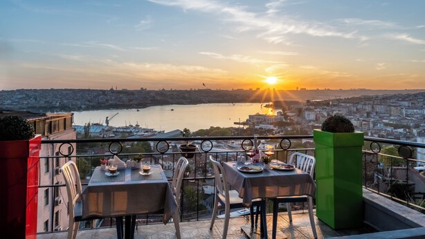 A terrace with tables and chairs overlooking a cityscape and a body of water during sunset. -  BİG URBAN STAY TAKSİM@WİTH SEA VİEW (Istanbul)