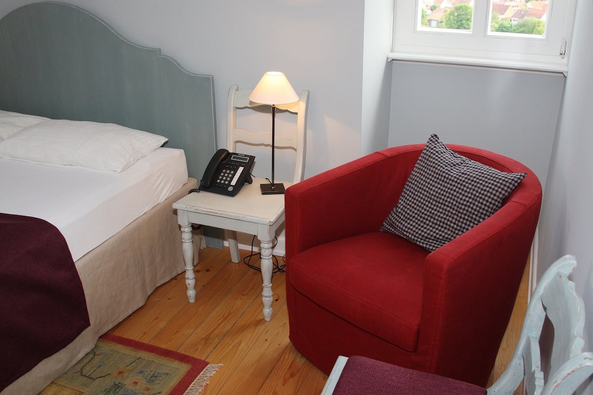 Una habitación con cama, una silla roja, una mesita con lámpara y un teléfono.