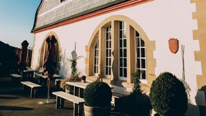 Un bâtiment doté d’un toit à forte pente, de fenêtres en arc de cercle et d’une façade en pierre. On y trouve des tables à l’extérieur et des plantes en pot devant l’entrée.