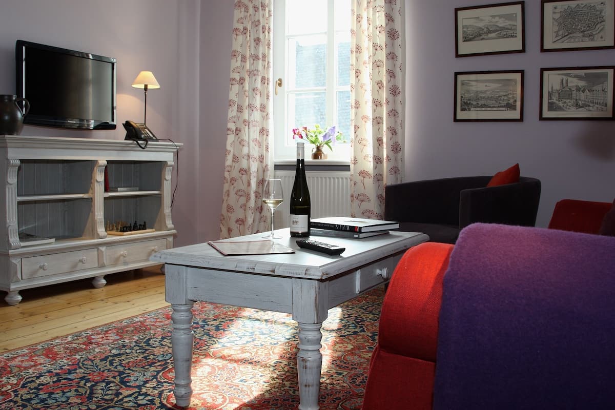 Una habitación con televisión, una mesa con una botella y un vaso, una estantería, una ventana con cortinas y cuadros enmarcados en la pared.