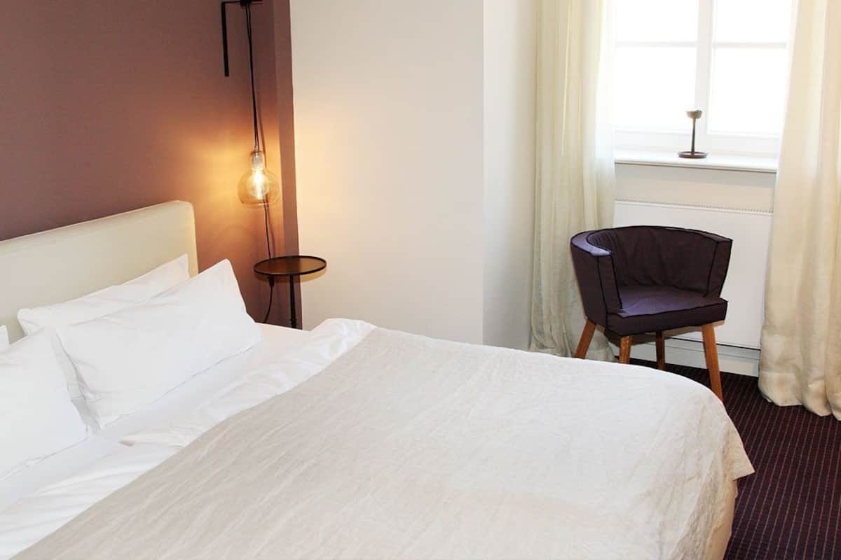 Una cama bien hecha con sábanas blancas, una mesita de noche con lámpara y una silla cerca de una ventana con cortinas.