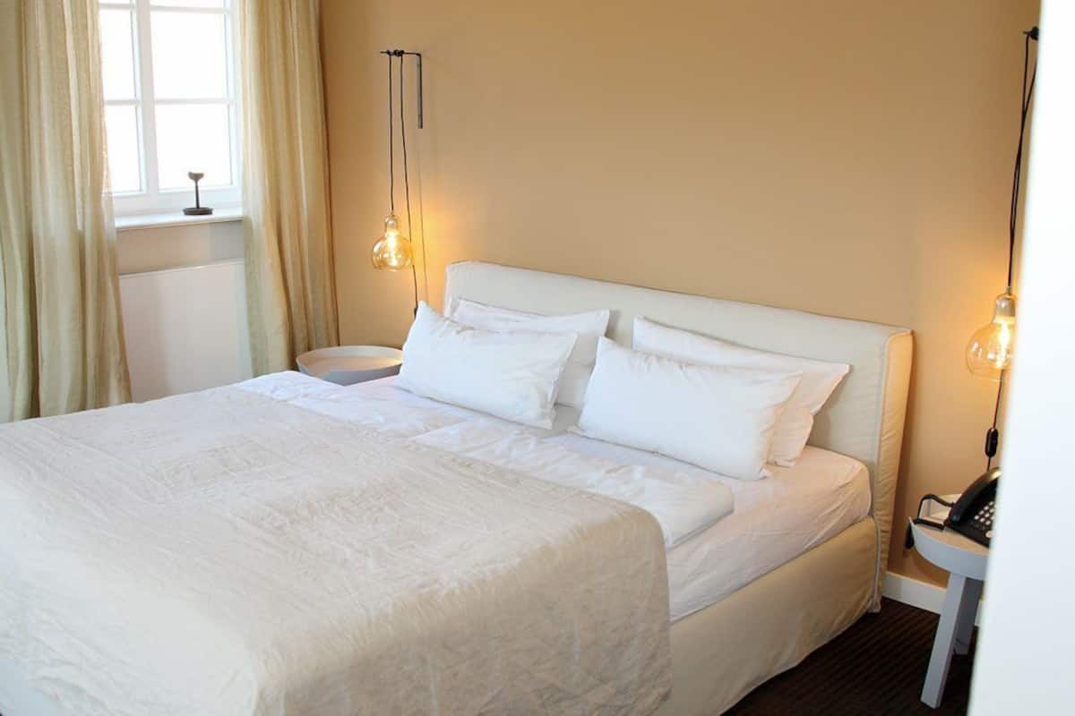 Una cama bien hecha con ropa blanca, una mesita de noche con lámpara y una ventana con cortinas.